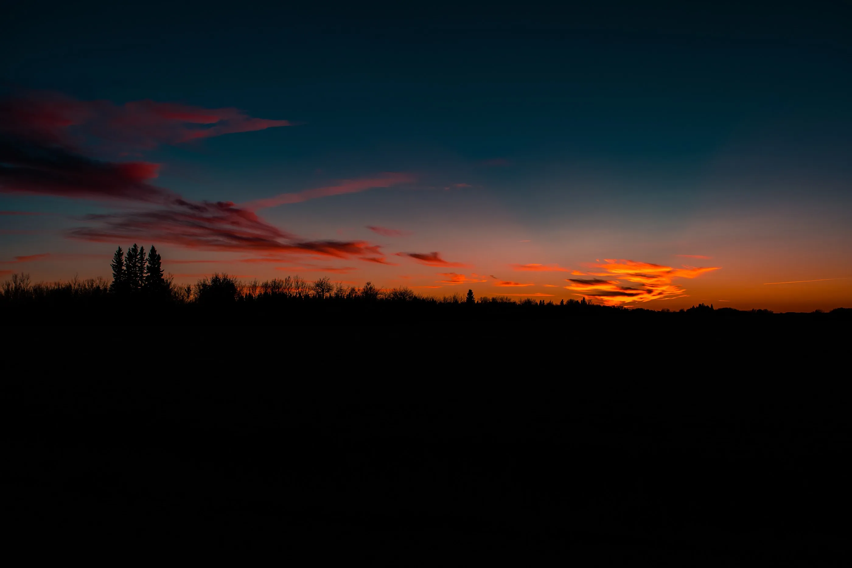 Orange sunset beneath dark clouds over a silhouetted treeline