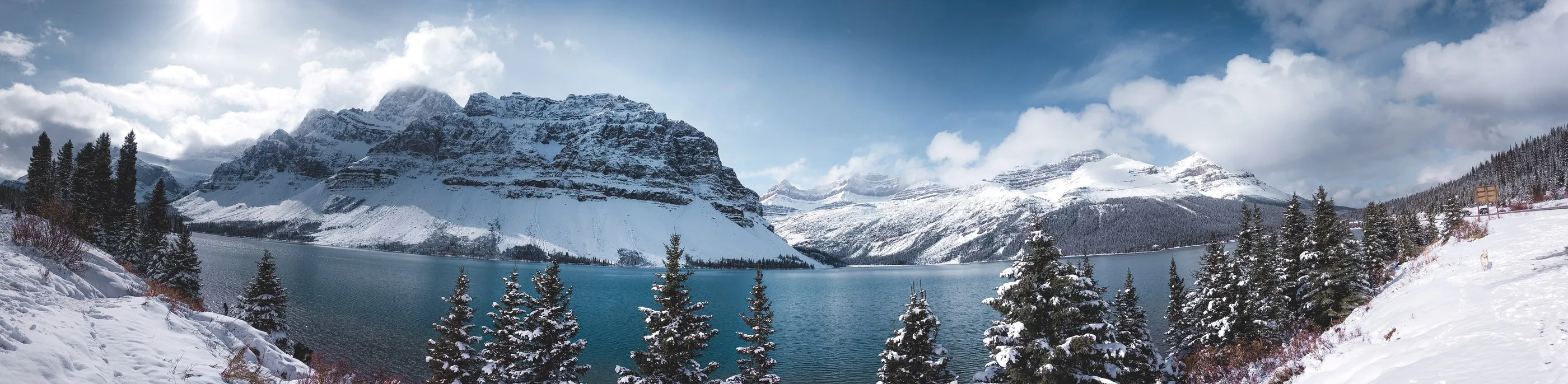 A panoramic view of turquoise water, snowy peaks, and evergreen trees